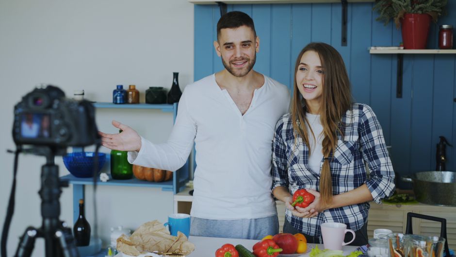 A cheerful couple records a cooking vlog in their modern kitchen, showcasing fresh produce.