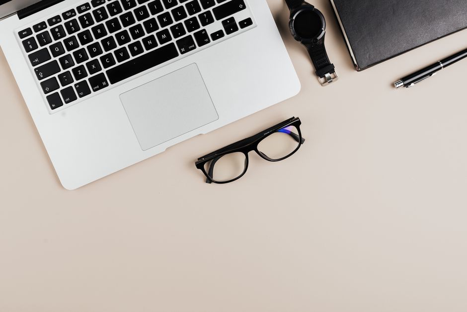 Modern workspace flat lay with laptop, notebook, watch, and glasses on a beige background.