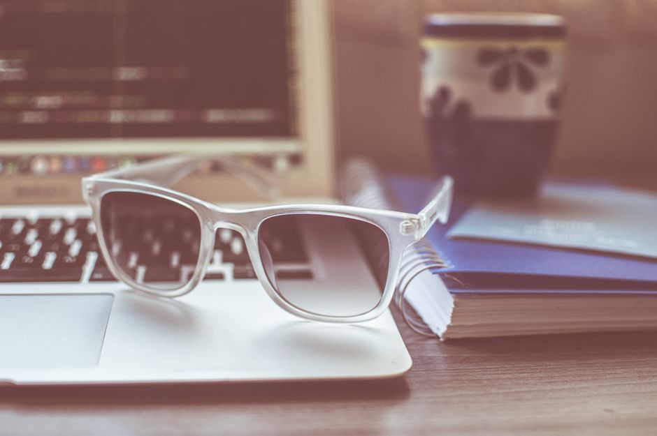 Close-up of sunglasses on a laptop, next to a mug and notebook. Ideal for work and lifestyle themes.