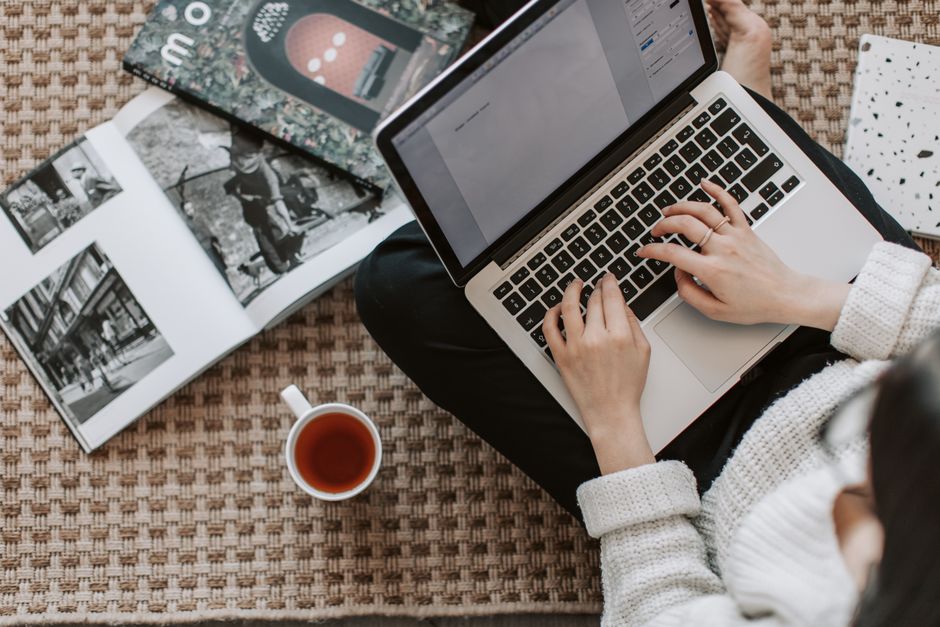 Top view of anonymous female freelancer in warm sweater drinking tea from white ceramic cup while sitting on floor carpet with netbook on legs near magazines and notebook and creating document for remote work project