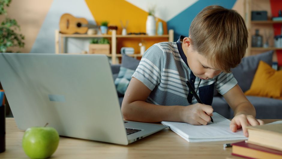 Young boy writing notes at home using a laptop for learning.