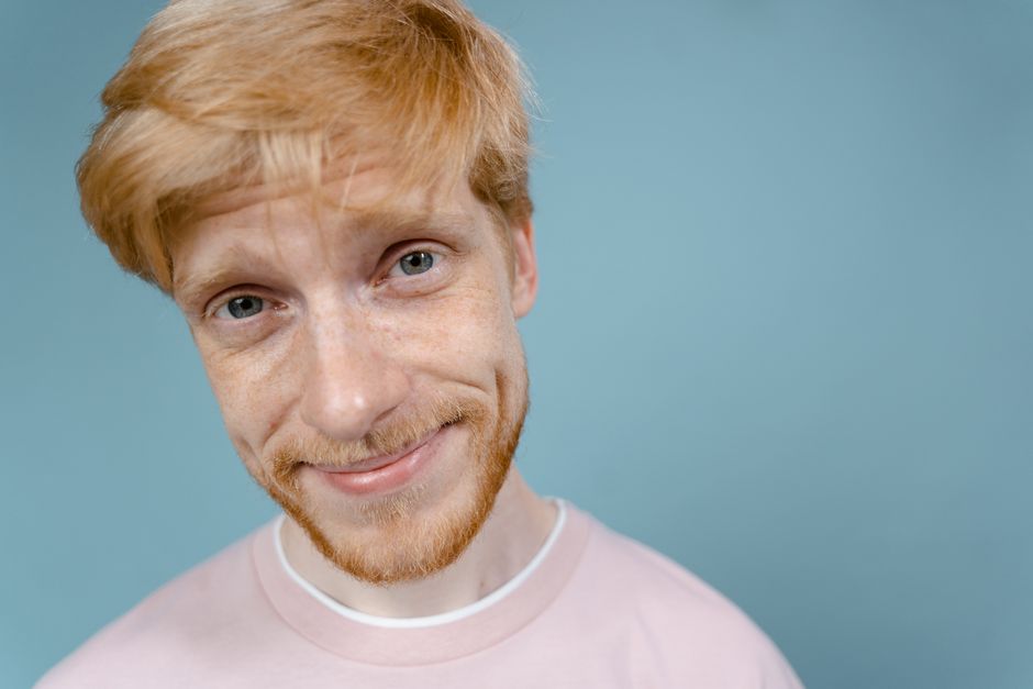 Cheerful bearded man with blonde hair smiling against a light blue background.