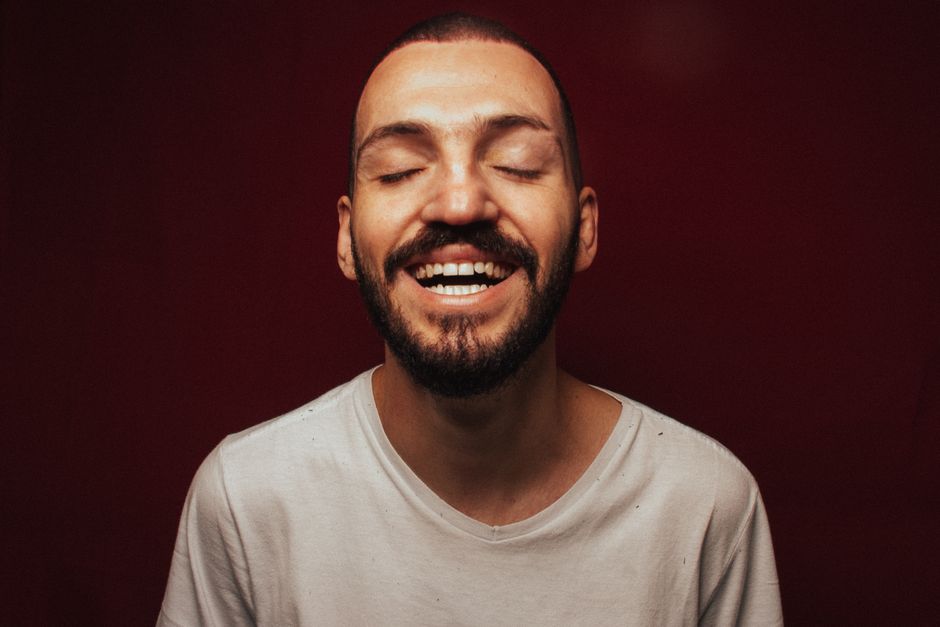 A happy, bearded man in a white t-shirt laughing with eyes closed against a red background.