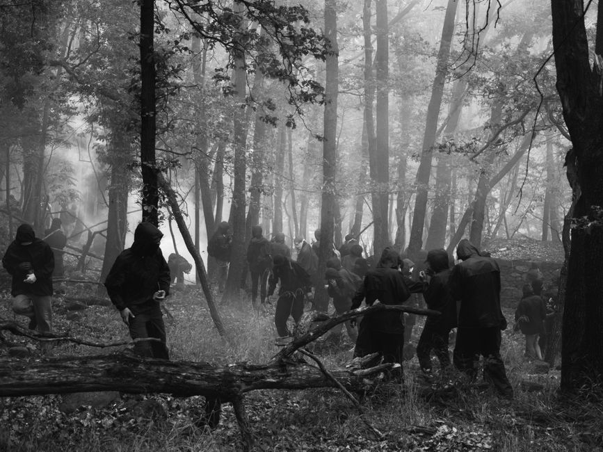 Black and white photo of hooded figures moving through a misty Chiomonte forest.