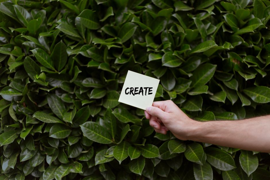 A hand holding a note with 'Create' written on it against a lush green leafy background.