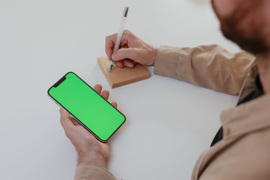 A person writing on a notepad while holding a smartphone with a green screen, ideal for mockups.