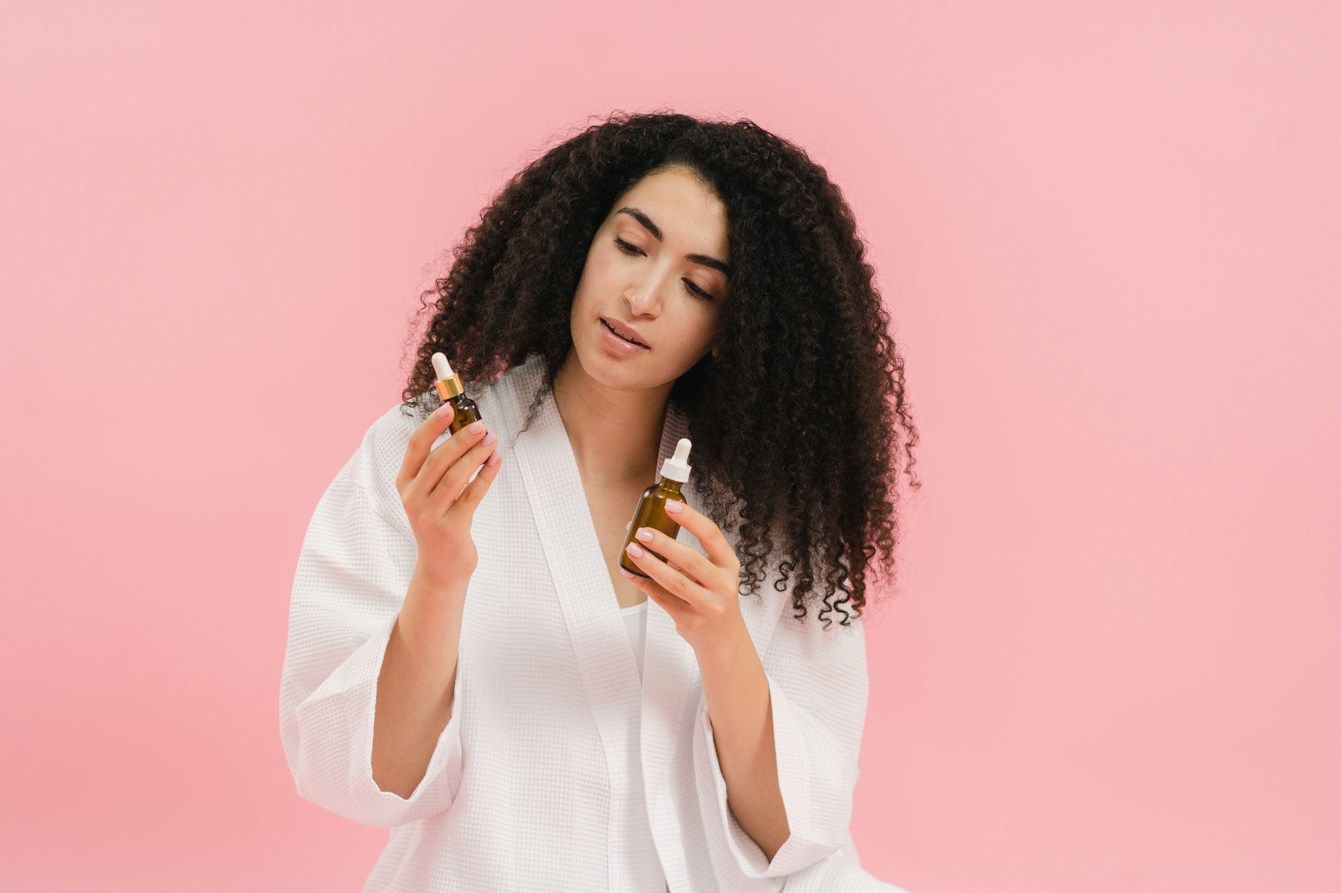 A woman with curly hair in a white robe examines two cosmetic oil bottles against a pink background.