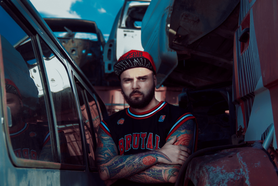 Man with tattoos and red cap in an urban junkyard, showcasing industrial style.