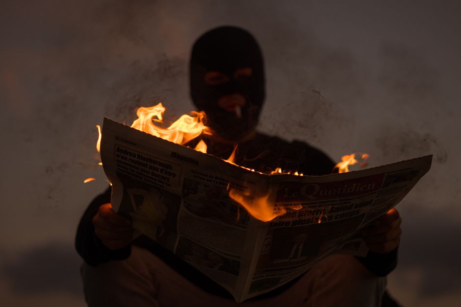 A masked person holds a burning newspaper in dramatic low light, evoking mystery.