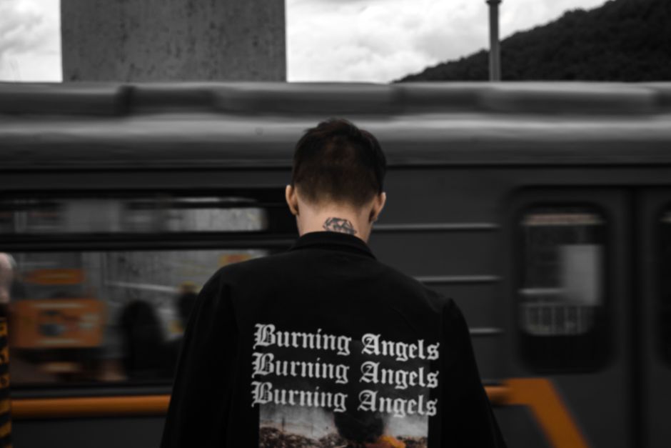 Man in a 'Burning Angels' jacket standing by a moving train in an urban setting.