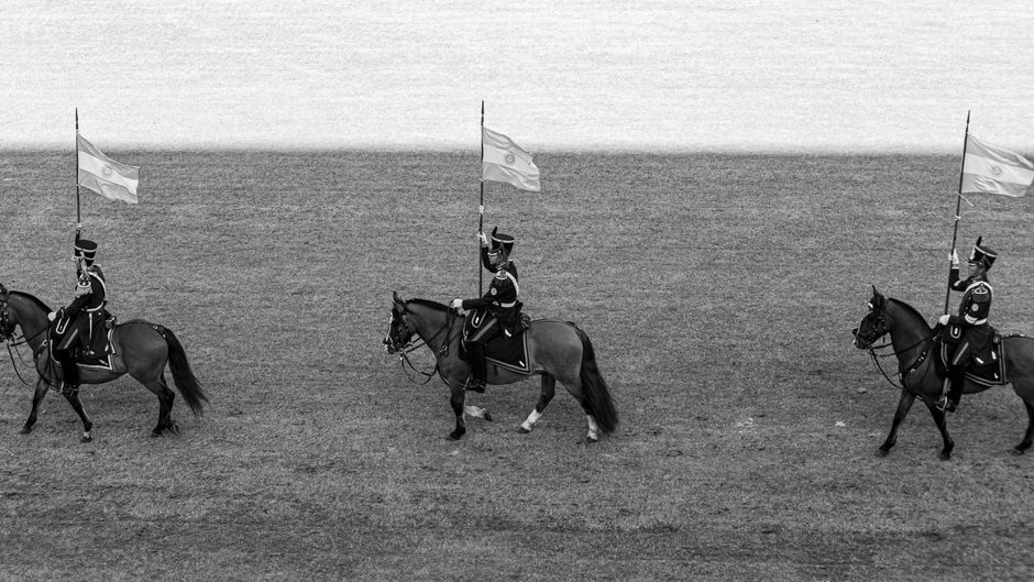 Three uniformed soldiers on horses carrying flags in a parade formation.
