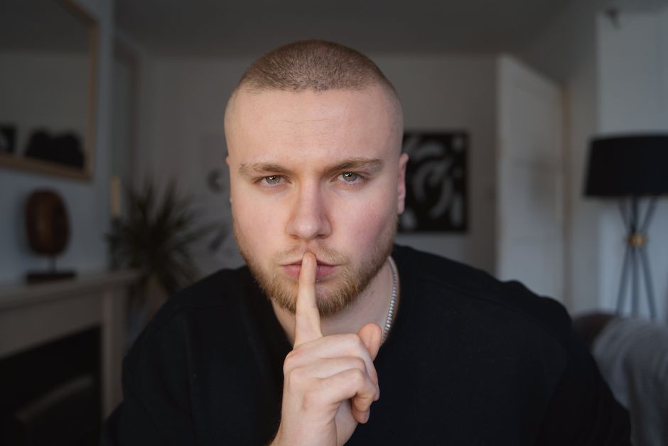 Portrait of a bald man with beard in a room, gesturing for silence. Mysterious expression.