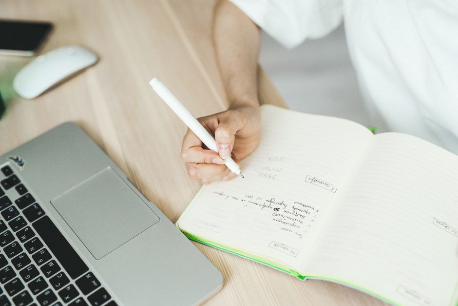 A hand writing in a notebook near a laptop, representing planning and freelancing.