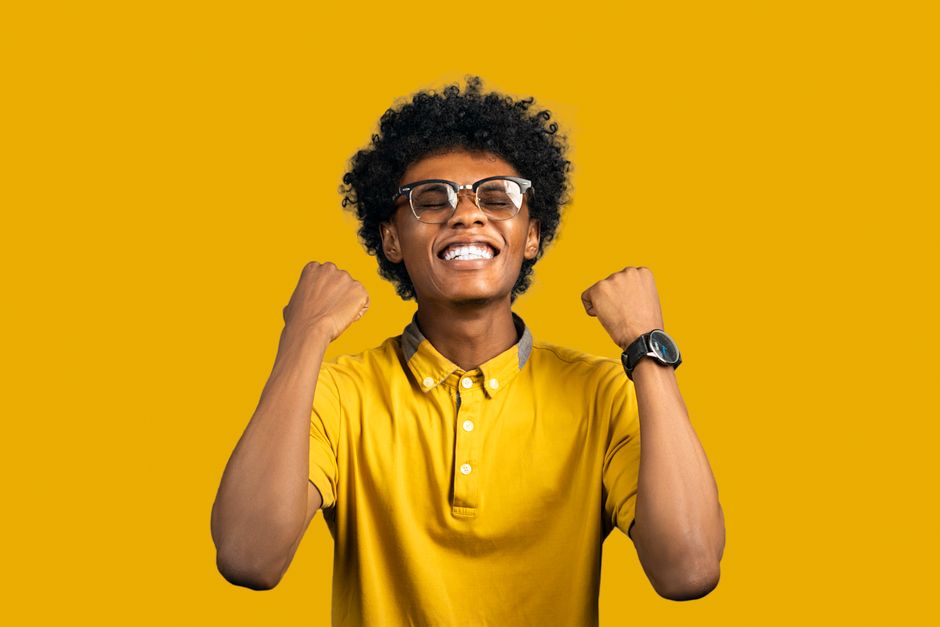 Happy young black male in bright t shirt and eyeglasses with Afro hairstyle standing with closed eyes while demonstrating achievement gesture on yellow background