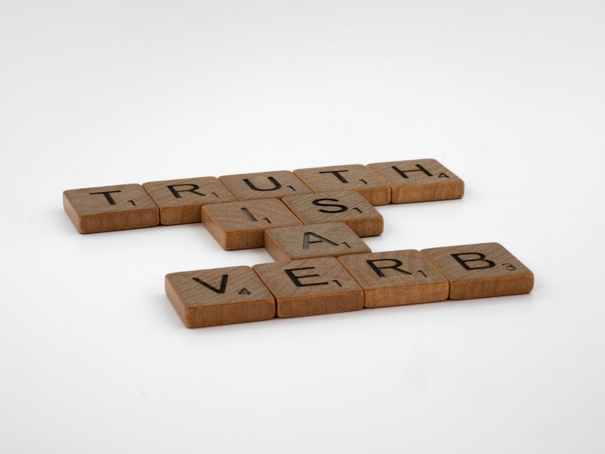 Close-up of wooden Scrabble tiles on a white surface spelling out 'Truth is a Verb'.